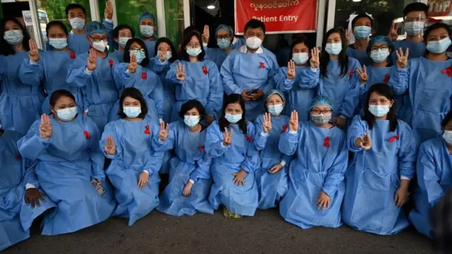 Nhân viên y tế dùng kiểu chào bằng ba ngón tay với dải ruy băng đỏ tại Bệnh viện Đa khoa Yangon vào ngày 3 tháng 2 năm 2021 hưởng ứng lời kêu gọi bất tuân sau cuộc đảo chính quân sự bắt giữ nhà lãnh đạo dân sự Aung San Suu Kyi