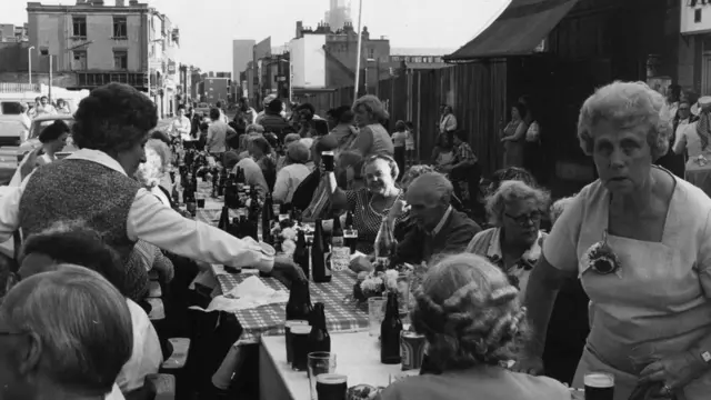 People gathered around a long table set with bottles and food for a street party for the Queen's Jubilee in 1977