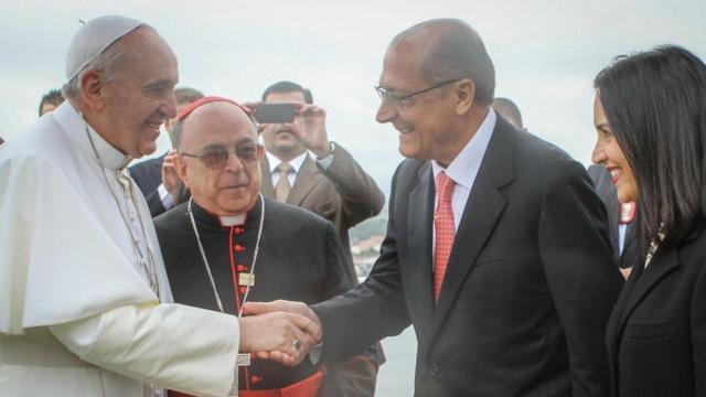 Fotografia colorida mostra o papa Francisco, um idoso vestido de branco, dando a mão para Geraldo Alckmin, um homem branco de óculos e terno