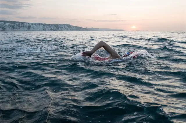 A woman has an early morning swim in the sea with cliffs behind her