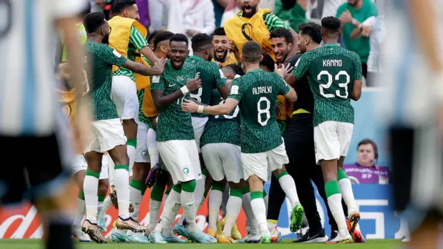 Fotografia colorida mostra jogadores da Arábia Saudita, usando um uniforme verde e branco, abraçados e comemorando