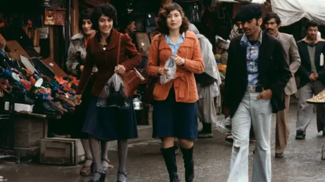 Mulheres com roupas típicas dos anos 1970 caminham pelas ruas, sem usar lenços