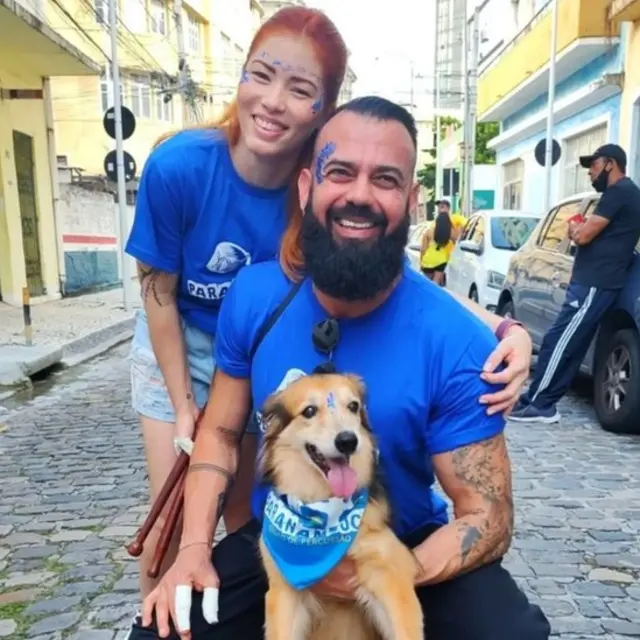 Fotografia colorida mostra um homem e uma mulher com um vira-lata caramelo