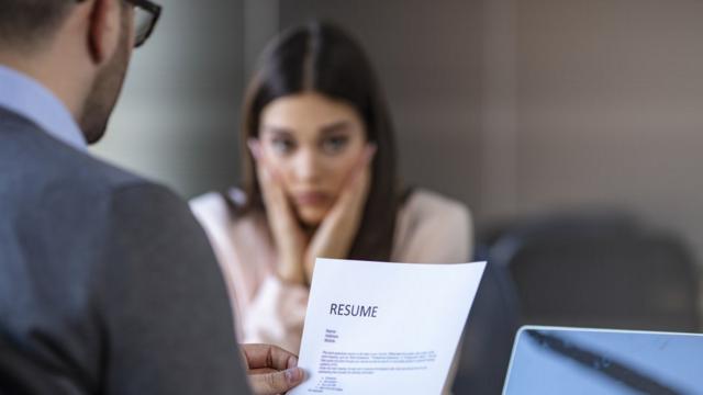 A young woman in an office looks worried while a man reads her resume