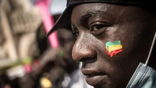Un manifestant observe la foule avec un drapeau malien peint sur la joue, lors d'une manifestation de masse à Bamako, le 14 janvier 2022, pour protester contre les sanctions imposées au Mali et à la junte par la Communauté économique des États d'Afrique de l'Ouest (CEDEAO)