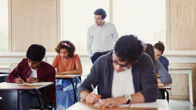Un maestro inspecciona a unos alumnos tomando un examen en un salón de clases