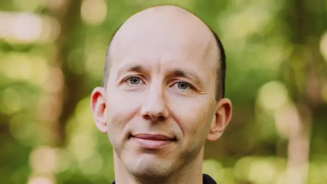 Fotografia colirda mostra um homem branco e careca sorrindo; ao fundo há o que parecem ser plantas