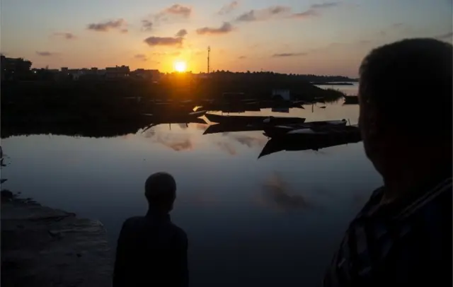 Des hommes se tiennent près des bateaux alors que le soleil est bas dans le ciel dans le district d'Al Maadea du gouvernorat de Behera.