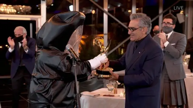 Юджин Леви получает статуэтку из рук "вручателя" в полной защитной экипировке