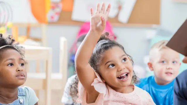 Imagem mostra menina com o braço levantado junto a outras crianças na escola