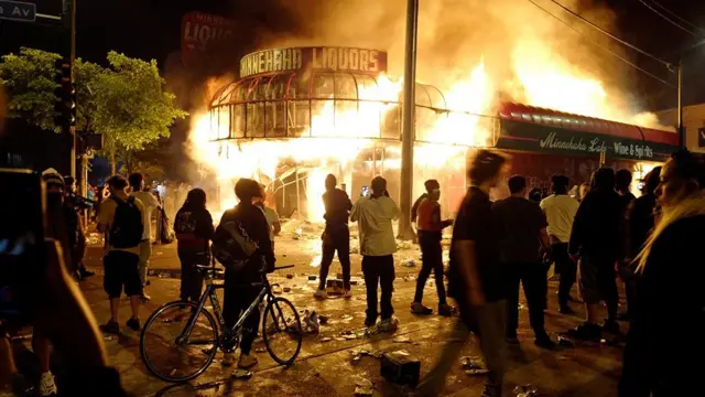 تظاهرکنندگان در مقابل یک مشروب فروشی که در آتش می سوزد