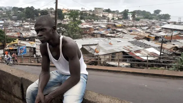 Un hombre no identificado aparece sentado en una colina de Freetown, rodeado de casas improvisadas de hojalata que se extienden hasta la orilla del mar.