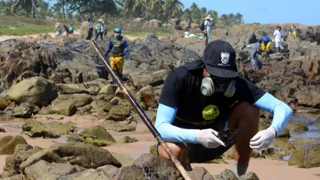 Com máscara e roupa com proteção UV, o voluntário Lucas Nelli agachado sobre pedras observando objeto com óleo; no plano2betsfundo, outros voluntários trabalham na praia