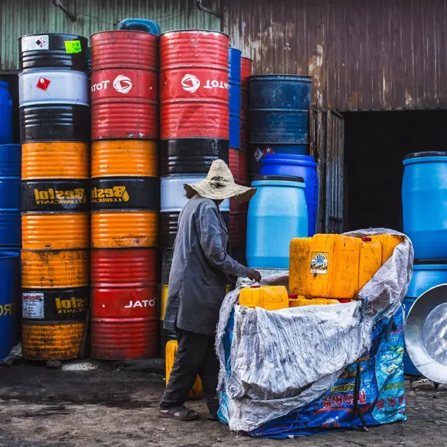 በሥርዓት የተደረደሩ በርሜሎች እና ጀሪካኖችን ከአንድ ሰራተኛ ጋር የሚያሳይ ምስል።