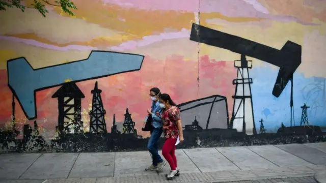 Duas mulheres caminham pela rua na Venezuela, ao ladobetpix originalum murobetpix originalque estão pintadas sondasbetpix originalextraçãobetpix originalpetróleo