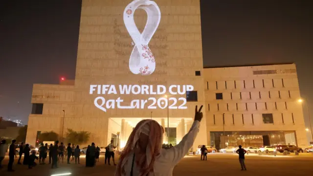 Homem com lenço usado comumente na cultura árabebet z365frente a um prédio com a sinalização da Copa do Mundo