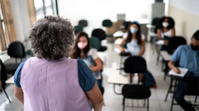 Professorafrases de aposta esportivapé efrases de aposta esportivacostas para a camera, olhando para alunos sentadosfrases de aposta esportivacarteiras