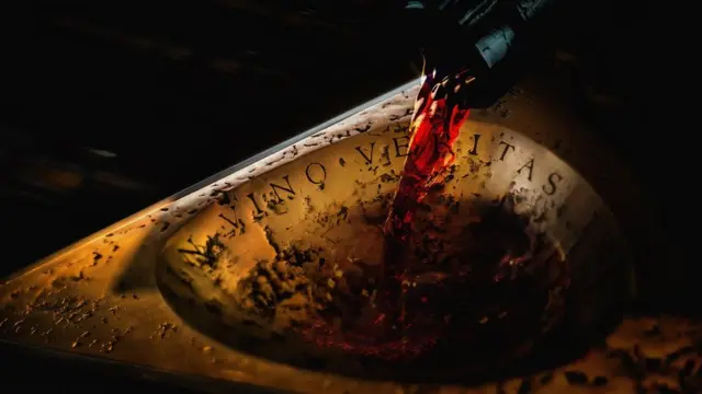 Dark bottle neck, pouring red wine into an ancient golden plate with the inscription "in vino veritas"