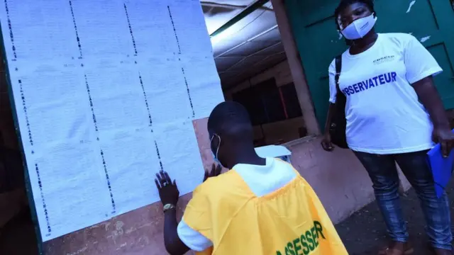 Un fonctionnaire de la Commission électorale nationale autonome (CENA) colle la liste des électeurs sur un mur avant l'ouverture des bureaux de vote, sous le regard d'un observateur électoral dans un bureau de vote, lors de l'élection présidentielle au Bénin, à Cotonou, le 11 avril 2021.