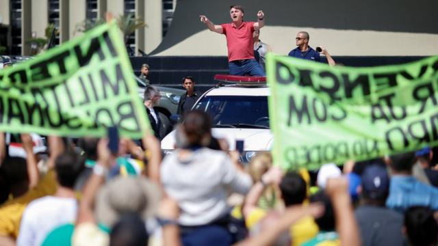 Bolsonaro discursa em cima de carro, observado por manifestantes que seguram faixas pedindo intervenção militar