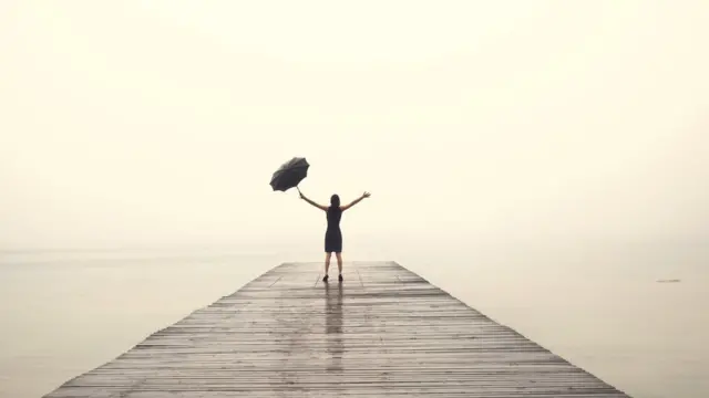 A woman wearing an elegant dress rejoices at the end of a pier, with their arms up in the rain and holding an umbrella