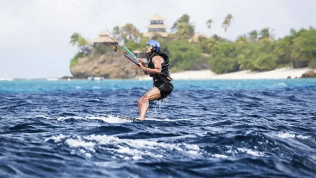 El ex presidente Barack Obama haciendo Kitesurf en el mar. Detrás de él una playa y palmeras.