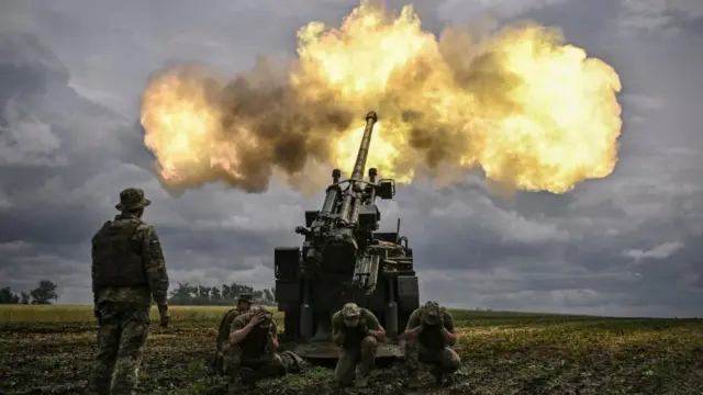 Fotografia colorida mostra homens com uniforme militar operando um canhão militar que solta fogoestrela bet contatoum campo verde