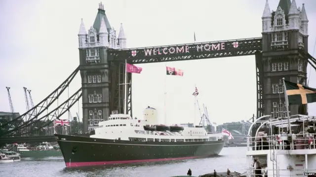Britannia проходит под Тауэрским мостом, на котором вывешено приветствие: "Добро пожаловать домой"
