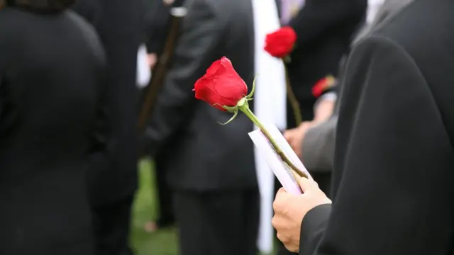 Pessoas com roupas pretas em pé em funeral, algumas segurando rosas