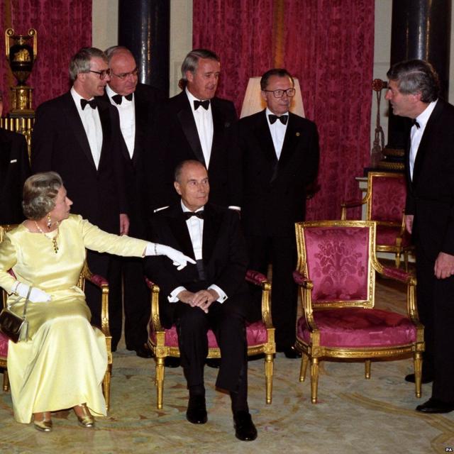 Elizabeth 2ª gesticula para Ruud Lubbers, primeiro-ministro da Holanda e presidente do Conselho de Ministros da Comissão Europeia, para se sentar em uma cadeira vazia enquanto os líderes dos países da cúpula do G7 se reuniam para uma foto antes do jantar no Palácio de Buckingham