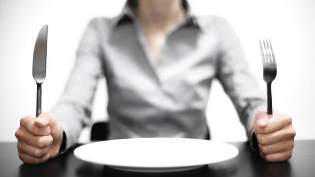 Mujer sentada frente a un plato vacío y empuñando un cuchillo y un tenedor