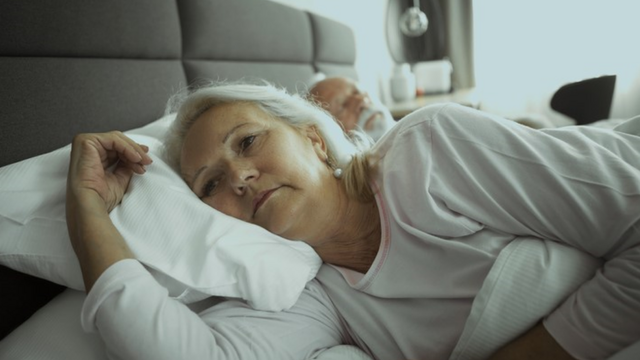 Fotografia colorida mostra um homem e uma mulher brancos idosos deitadosjogo de cartas estrela betuma cama; ele dorme, mas ela está acordada, com olhar perdido