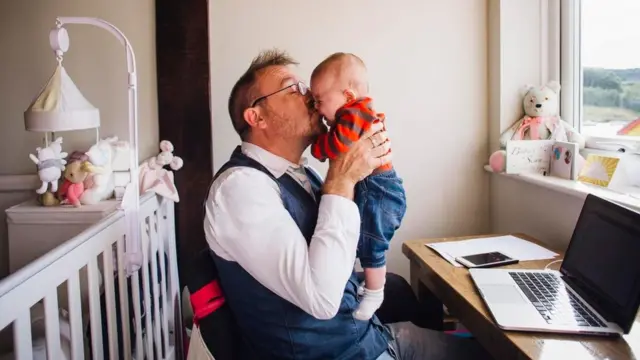 Un hombre sentado frente a su laptop, cargando y besando a un bebé