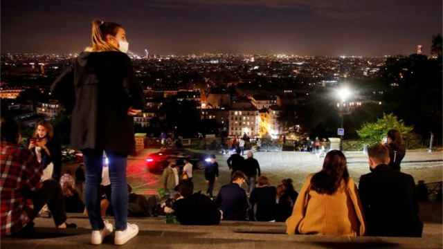 Diversos jovens sentados em escadaria com roupas de frio, à noite, diante de vista da parte baixa de Paris