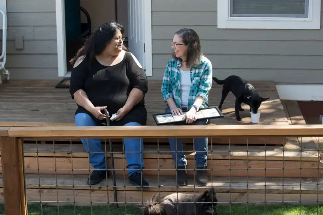 Sherry Mesa (E) sentada com Martha Chambers (D) e um cachorro ao lado, em frente à microcasa em que vive com a sobrinha nos EUA