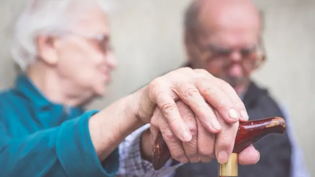 Dos ancianos sentados, él con bastón y ella cogiéndole una mano.