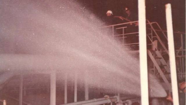 Foto antiga, mas colorida, mostra dois homenseleição pixbetparte superioreleição pixbetnavio usando mangueira para conter incêndio