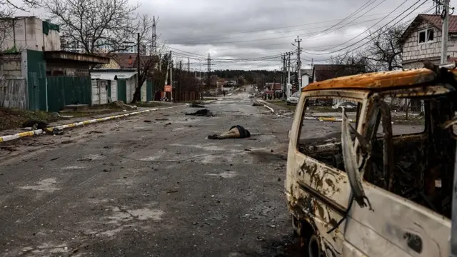 Rua com destroçosbullsbet site oficialveículos e prédios, sob um céu nublado. No chão, à distância, dois corposbullsbet site oficialpessoas caídos.