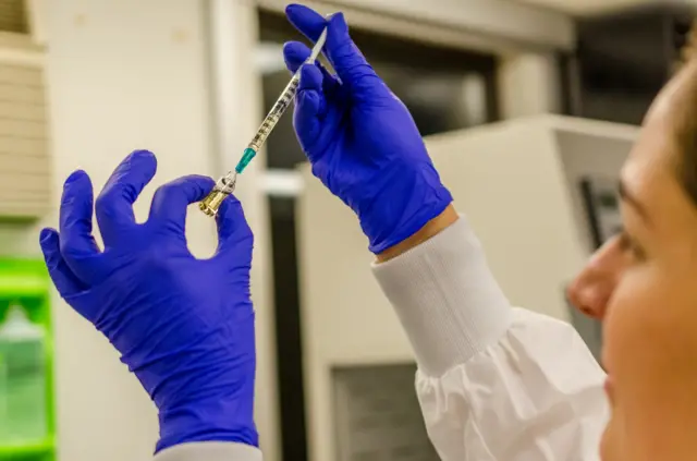 Una mujer realiza investigaciones en un laboratorio. Tiene en sus manos un jeringa y un recipiente.