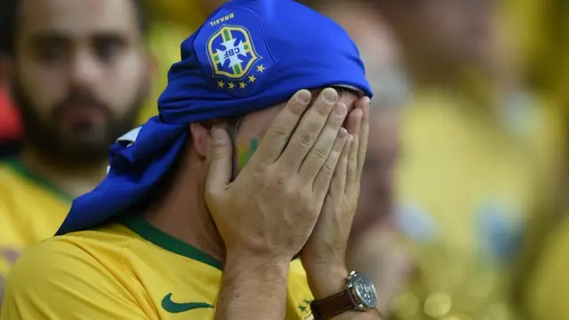 Torcedor brasileiro desolado, com as mãos cobrindo o rosto; ele veste camiseta da seleção verde e amarela e outra camisa da seleção azul na cabeça.