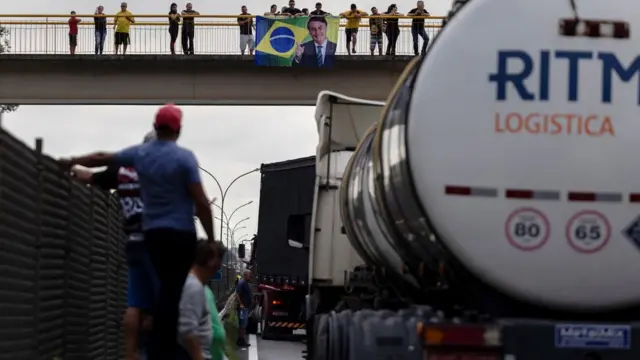 Caminhoneiros e caminhões paradosvbet deestrada; ao fundo,vbet depassarela, apoiadores do presidente seguram bandeira com fotovbet deBolsonaro