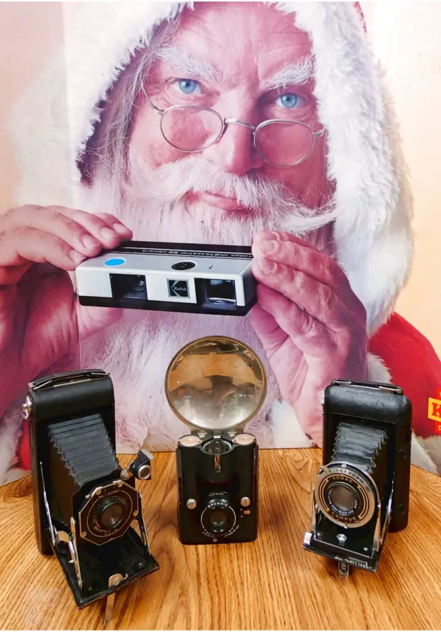 U pozadini: reklama za foto-aparat Kodak instamatik. U prvom planu, sleda nadesno: Broj jedan Kodak brauni plajant, Šest-16 aparat na razvlačenje (SAD); Braunijev blic, Šest-20, c.1941; Kodak ridžent, blenda Kompur, sočivo Zajs Tesar, aparat na razvlačenje, c.1935-39 (Nemačka)