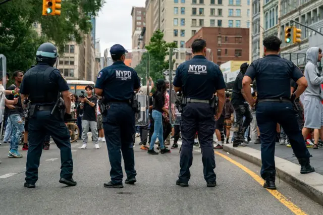 Four police officers have their backs to us as they provide security in Union Square as popular live streamer, not shown, stages a giveaway. They have NYPD on their shirts and one of them wears a helmet. 