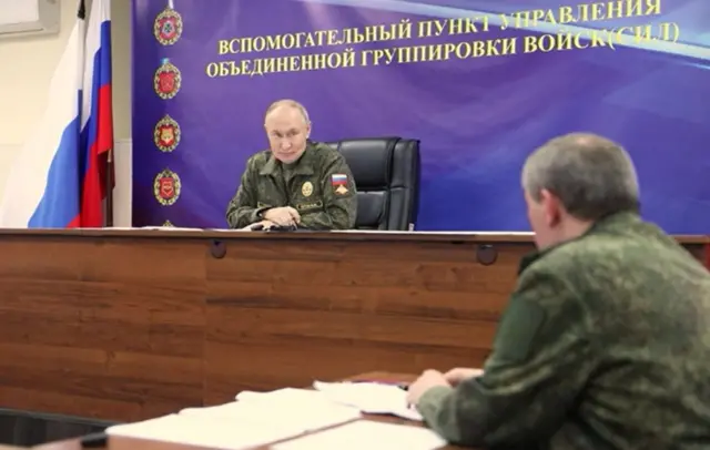 Владимир Путин сидит за большим столом и слушает Валерия Герасимова, который сидит спиной к камере