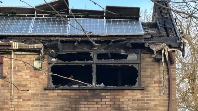 Primer plano del último piso de la casa de ladrillo. Una ventana rectangular está completamente destrozada y muestra la habitación ennegrecida del interior. El canalón sobre la ventana presenta daños por fuego; algunas partes parecen haberse derretido. El techo también está dañado y está cubierto de paneles solares; algunos se han desprendido.