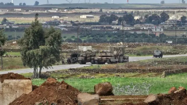 Несколько единиц военной техники на дороге, на переднем плане насыпанная земля, на заднем - дома