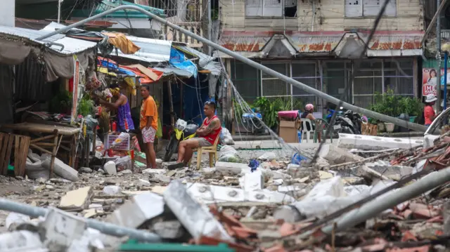 Человек сидит на пластиковом стуле посреди развалин, рядом стоят еще люди.