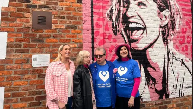 Un grupo de cuatro personas (tres mujeres y un hombre con Marie Harding vistiendo un abrigo negro) se encuentran frente a un gran mural de Sarah Harding.