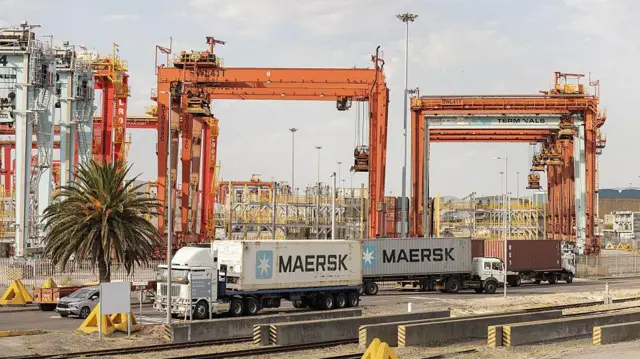 One busy container terminal show several tall orange crane wey rise over rows of shipping containers, with two Maersk-branded container trucks wey dey drive past for di background on one paved road beside railway tracks, and a single palm tree wey stand to one side.