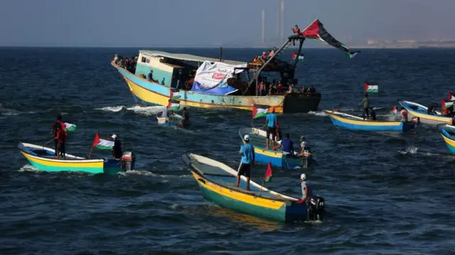 হলুদ ও সবুজ রঙের পাঁচটি ছোট কাঠের মোটরচালিত ডিঙ্গি একটি বড় মাছ ধরার নৌকার পাশে রয়েছে, সেটিও একই রঙের। নৌকাগুলোতে লাল, সাদা ও সবুজ রঙের ফিলিস্তিনি পতাকা উড়ছে। ছোট নৌকাগুলোতে একজন বা দুজন লোক দাঁড়িয়ে বা বসে আছে। বড় নৌকাটিতে কয়েকজন মানুষ রয়েছে। দূরে সমুদ্রের ওপারে উঁচু চিমনি ও শিল্প স্থাপনা দেখা যাচ্ছে।